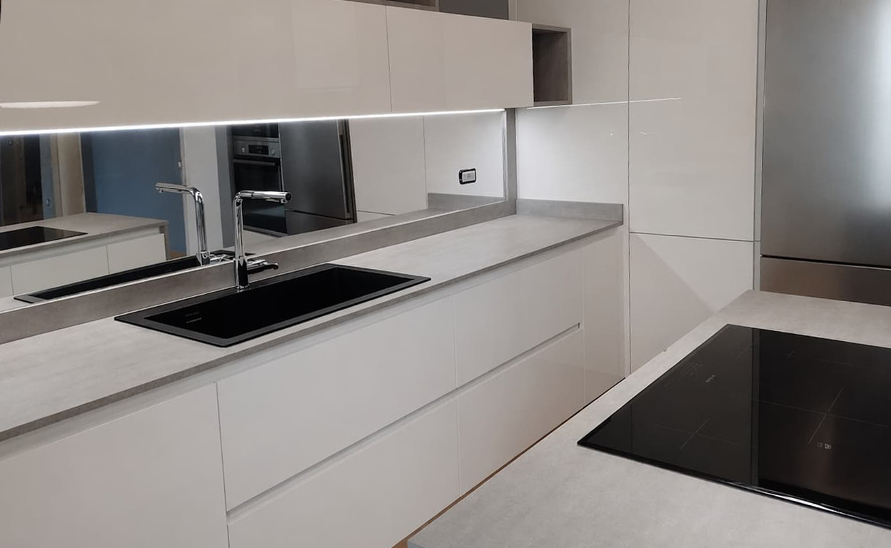 Cucina moderna bianca con piani in finitura chiara, due fuochi in vetro nero e dettagli in acciaio, ambiente minimale e luminoso