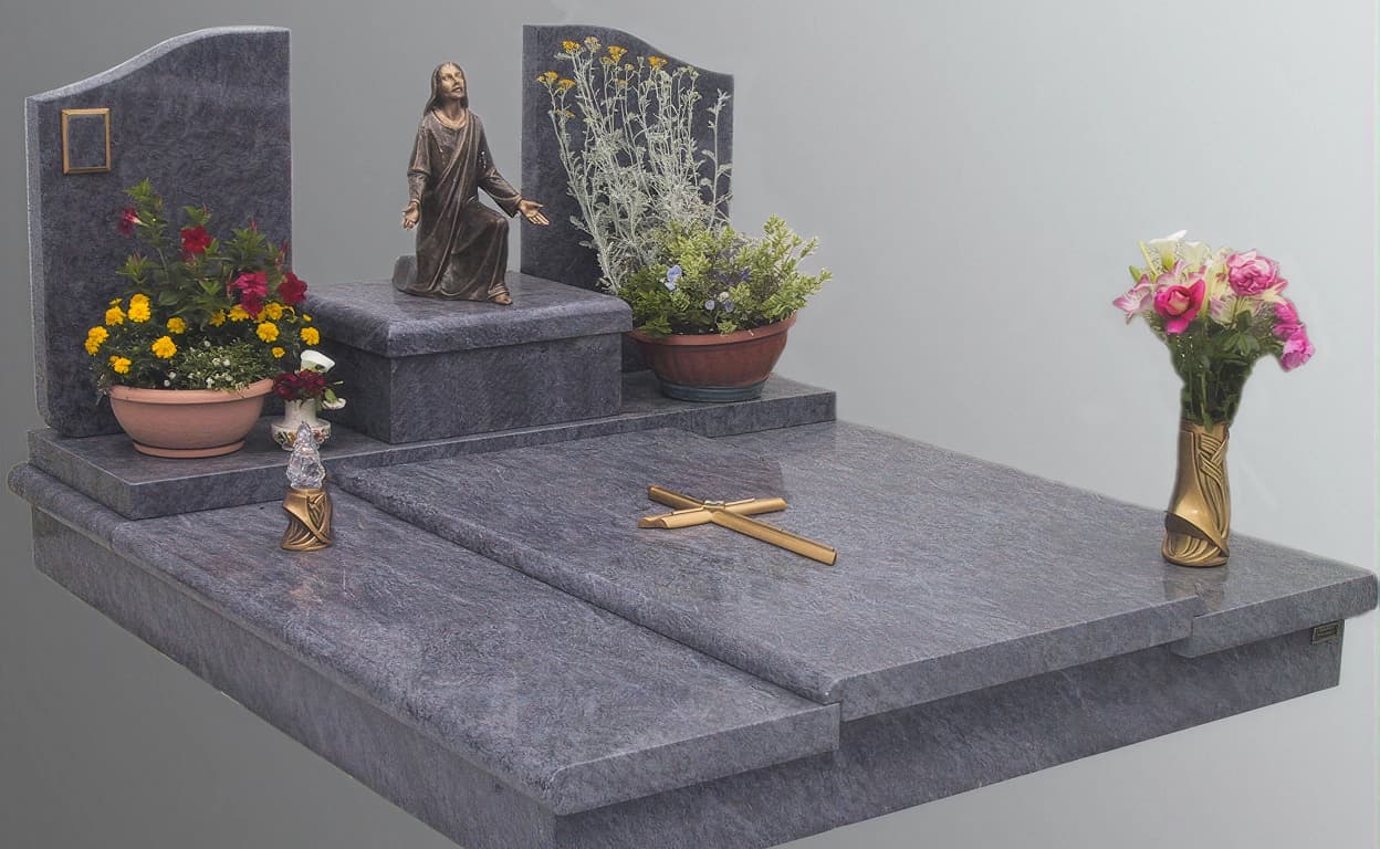 Tomba in marmo grigio con statua religiosa su lapide, fiori e candelabro, croce e crocifisso posizionati sul ripiano del mausoleo.