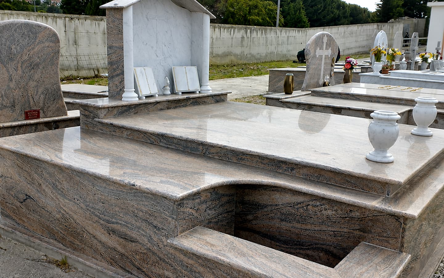 Vista ravvicinata di una tomba in marmo e granito in un cimitero, con lapidi, croci bianche, fiori e cuscinetti commemorativi su vialetti e prato.