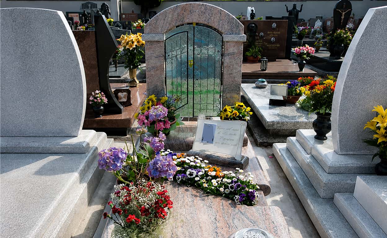 Cimitero con tombe in pietra grigia e lapidi circondate da fiori colorati e composizioni floreali, con sentiero centrale e cappelle sullo sfondo