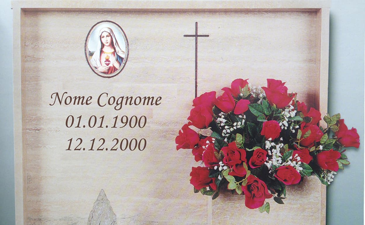 Lapide commemorativa con croce centrale, immagine religiosa, nome e date di nascita e morte, e un grande bouquet di rose rosse e fiori sul lato destro.