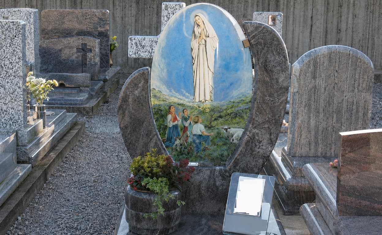 Cimitero con tomba decorata da bassorilievo religioso: immagine della Madonna su sfondo azzurro, croci e lapidi in granito, piante e fiori davanti