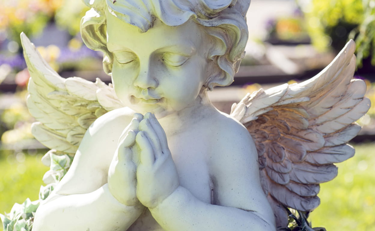 Statua angelica in marmo bianco con ali e aureola, figura in preghiera con le mani giunte e atmosfera luminosa in un giardino colorato.