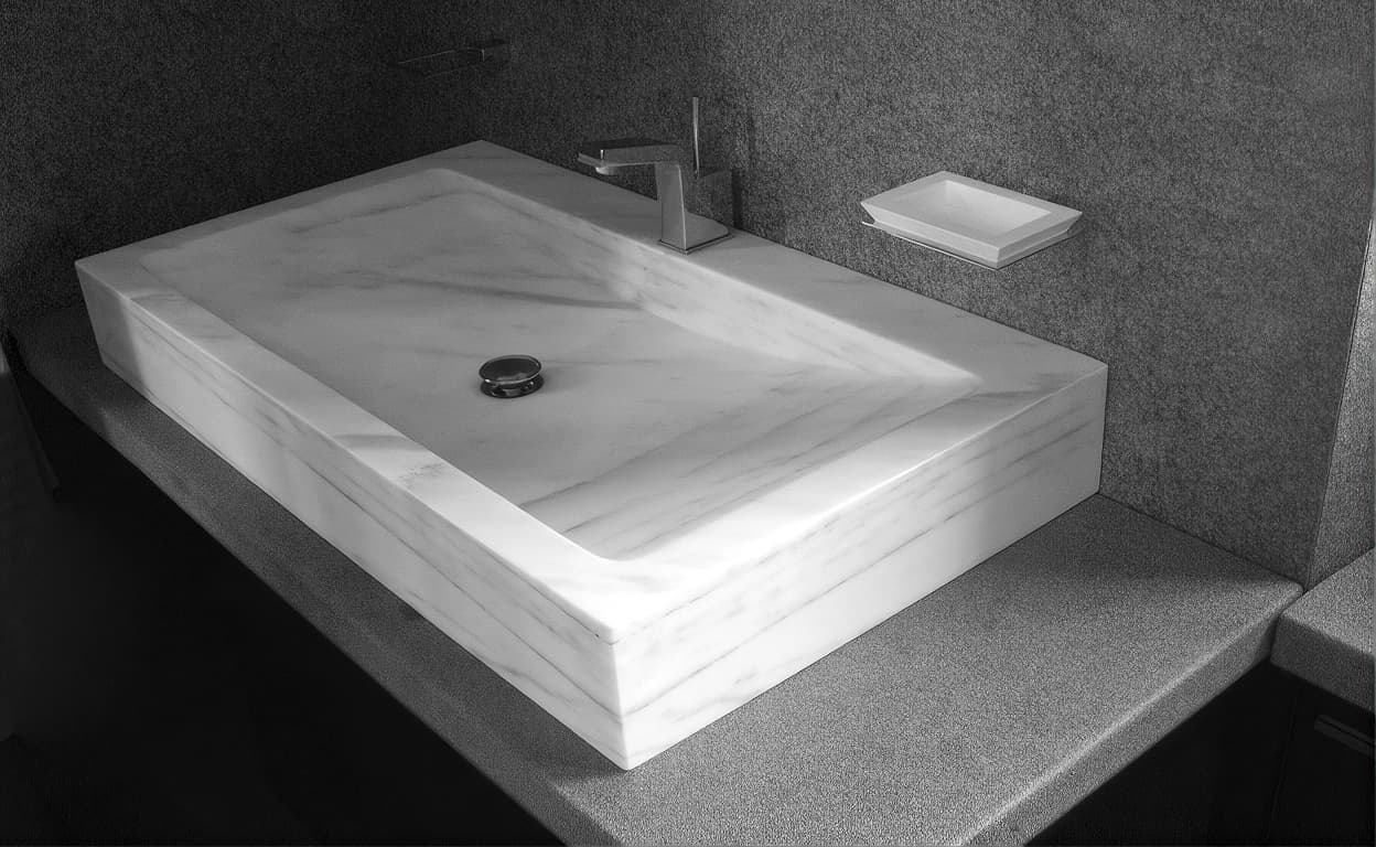 Coppia di lavabi in stile minimal, vasca da bagno rettangolare in pietra chiara su parete grigia, rubinetto e piano d’appoggio con luce soffusa.