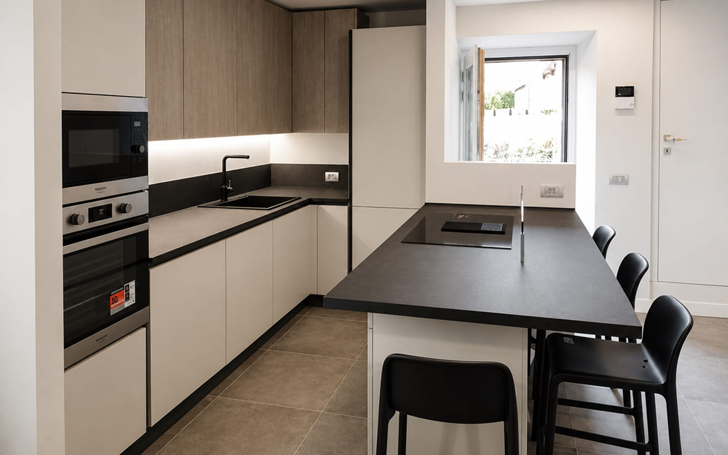 Cucina moderna in stile minimal con mobili bianchi e rovere, piano scuro, lavello in acciaio, forno e piano cottura integrati, tavolo con sedie e finestra luminosa.