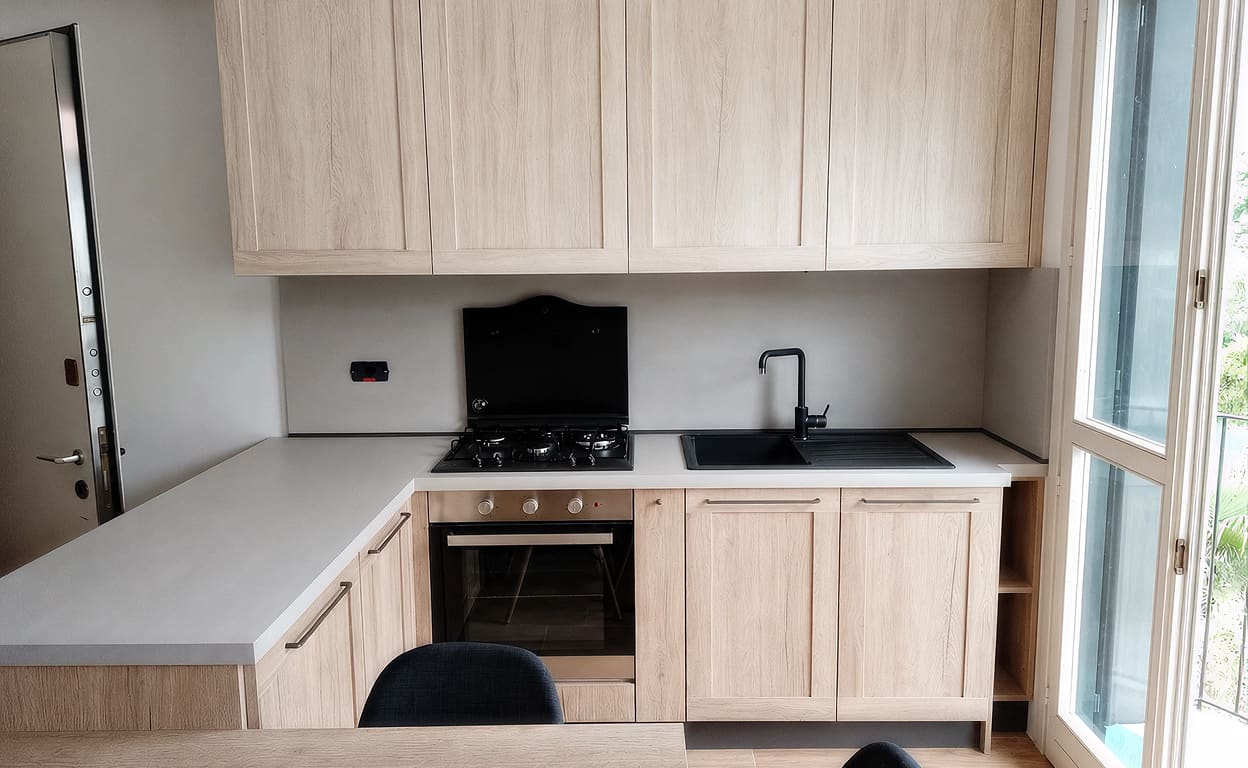 Cucina moderna con mobili in legno chiaro, piano bianco e fornello a gas incassato, forno e lavello con rubinetto nero, in ambiente luminoso vicino alla finestra