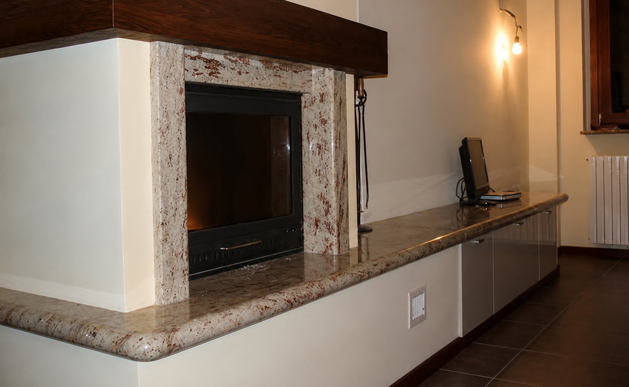 Interno di una casa con camino in pietra con insert in metallo, pareti bianche e legno scuro, mensola e cucina minimalista con luci calde.