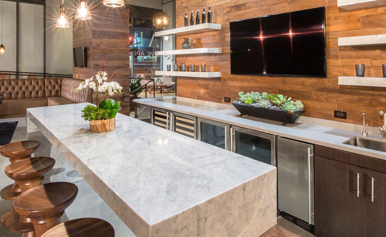 Cucina moderna con isola in marmo chiaro, lavello integrato, sedute in legno scuro e pareti in doghe, luci a sospensione e TV a schermo in soggiorno