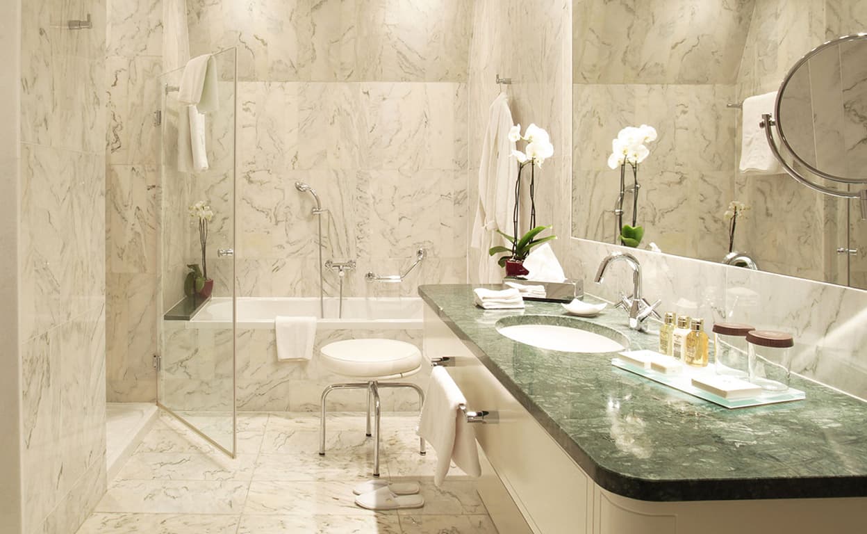 Bagno moderno con piatto doccia in vetro, vasca con ripiano in marmo verde, lavandino doppio e specchio rotondo, finiture in pietra chiara e luci soft