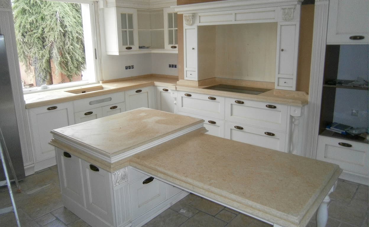 Cucina in stile classico bianca con ampio piano in pietra beige, cassetti e ante decorate, isola centrale e grande finestra: ambientazione luminosa e ordinata