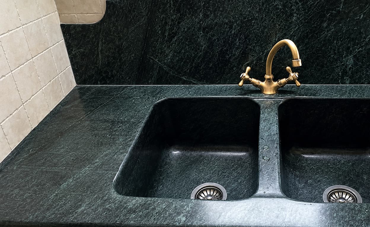 Lavello da cucina in pietra scura con due vasche rettangolari, scolapiatti integrato e rubinetto color ottone, dettaglio su fondo e scarico.