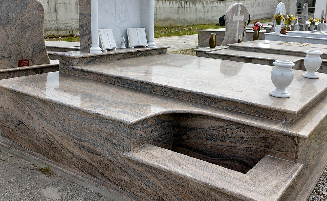 Primo piano di una tomba in marmo grigio in un cimitero, con lapidi e portafiori sullo sfondo, vista ravvicinata e dettagli della pietra